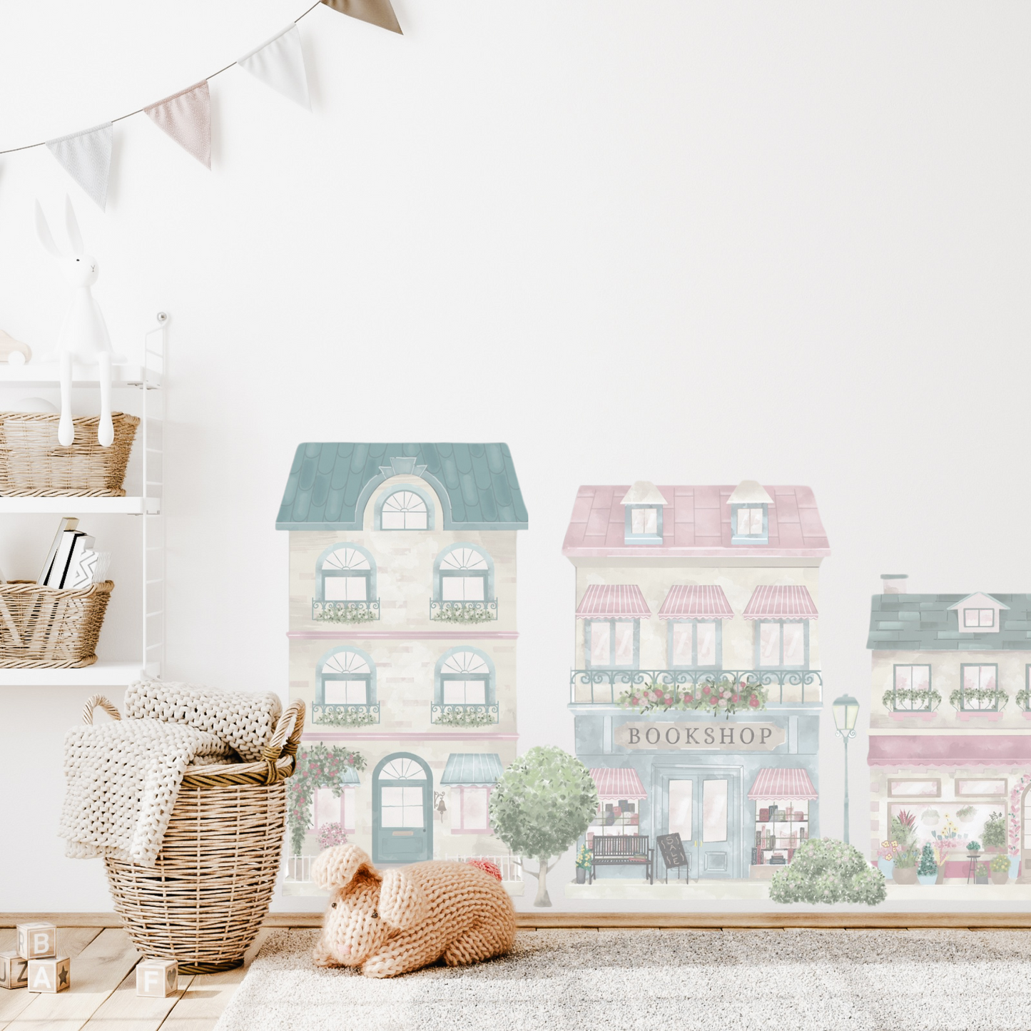 A wall decal set depicting a little village with colourful buildings, placed in a children's room setting with a basket and decorative flags in the background. The town features a house, café, florist, bakery, bookshop, dress shop as well as trees, bushes, a bike and ice-cream cart.  Non-toxic, eco-friendly, reusable wall stickers. Australian made.