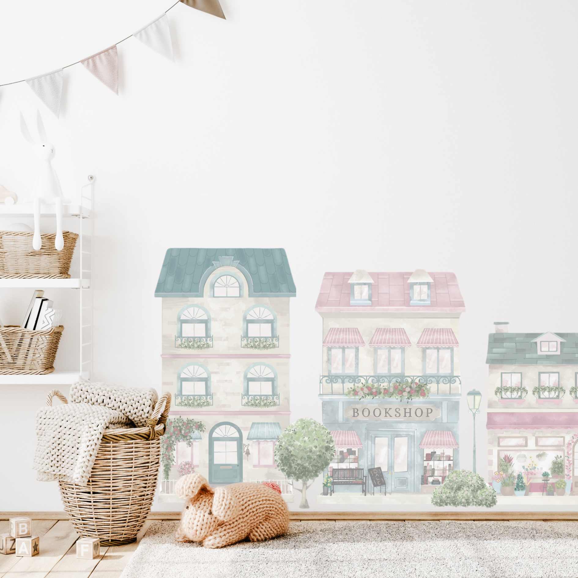 A wall decal set depicting a little village with colourful buildings, placed in a children's room setting with a basket and decorative flags in the background. The town features a house, café, florist, bakery, bookshop, dress shop as well as trees, bushes, a bike and ice-cream cart.  Non-toxic, eco-friendly, reusable wall stickers. Australian made.